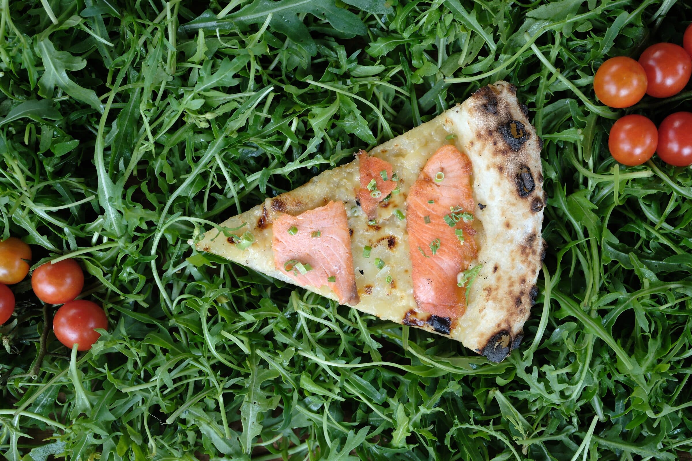 Miếng pizza cá hồi trên nền rau rocket và cà chua bi, viền bánh lấm tấm đốm cháy xém